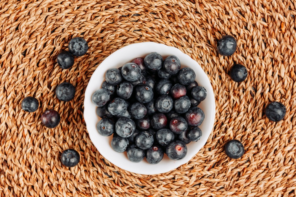 A white bowl filled with fresh blueberries sits on a woven, brown placemat, with a few berries scattered around the bowl.