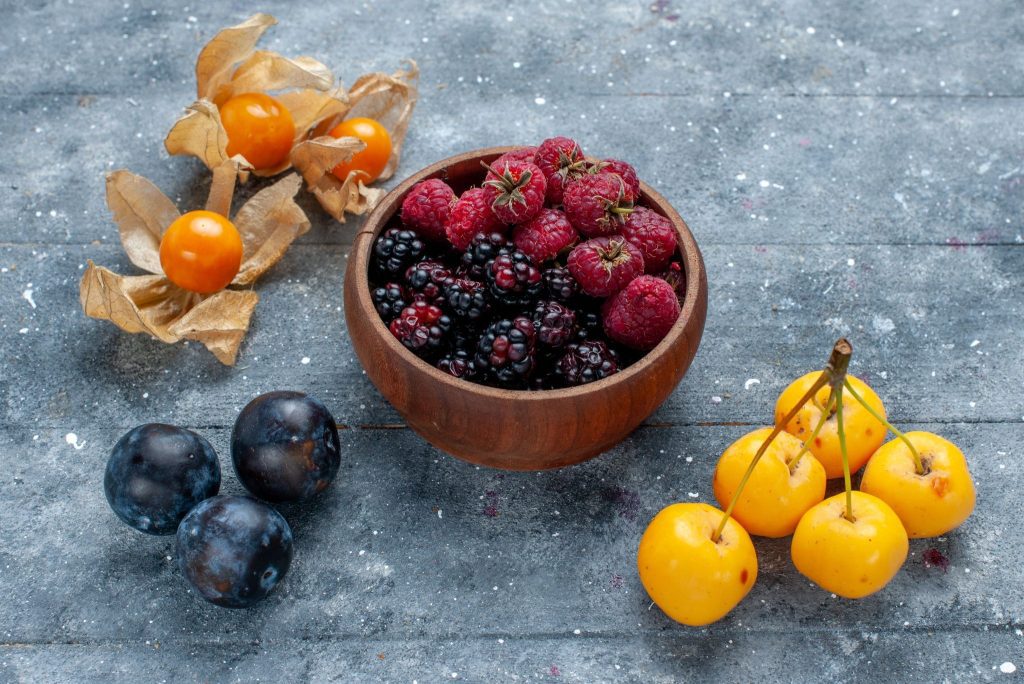 A wooden bowl with raspberries and blackberries, surrounded by yellow gooseberries, dark plums, and yellow cherries on a gray surface.