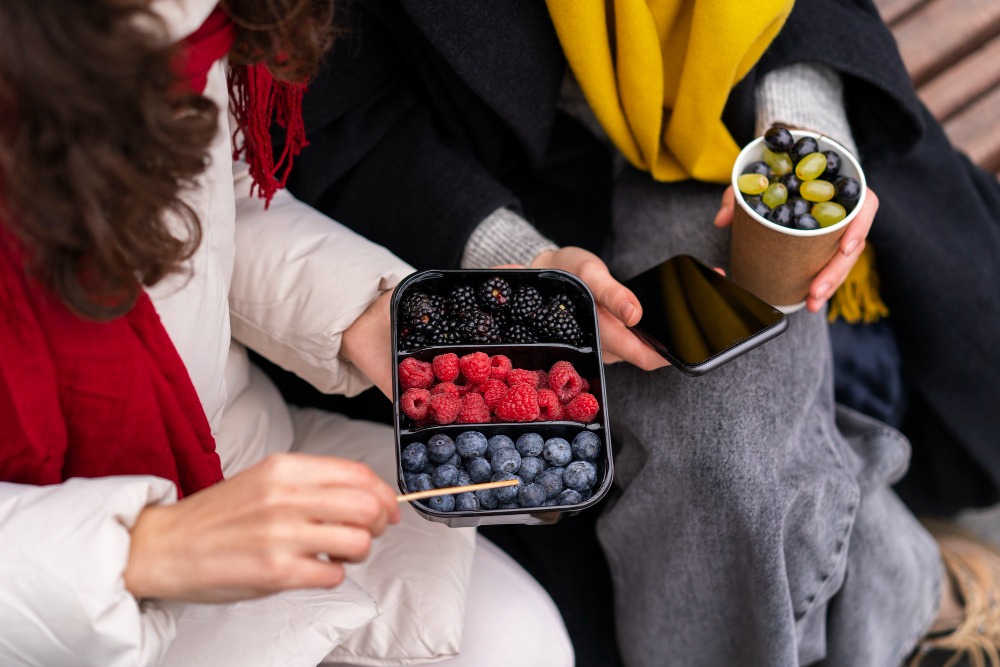 Two people sitting side by side, one holding a container with rows of blueberries, raspberries, and blackberries, the other holding a cup of grapes and a smartphone.